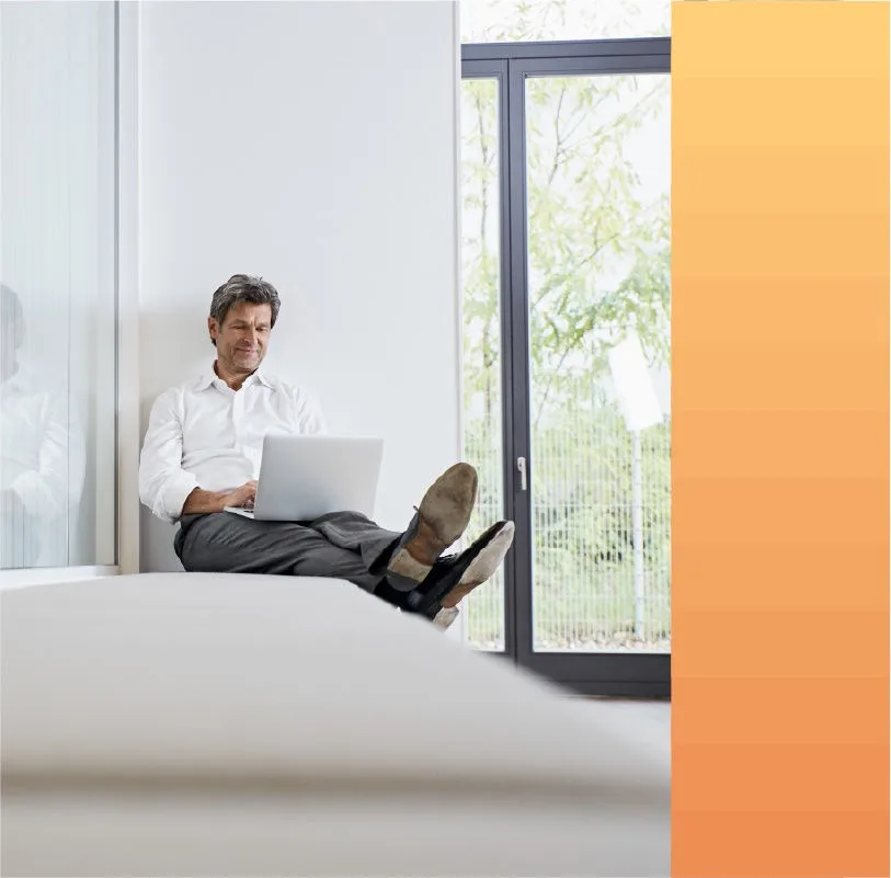 Man reclines on a bench while working from his laptop in an office building