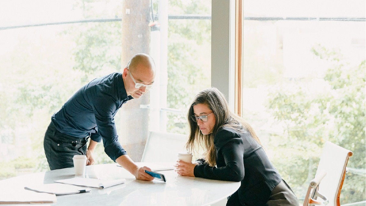 Two colleagues review information on a tablet at a table in a bright office.