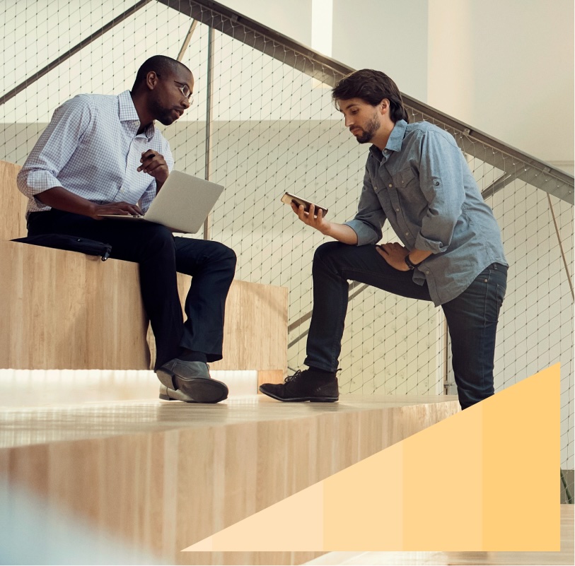 Two businessmen, one with a laptop and the other with a mobile phone, collaborate while sitting on a set of office auditorium steps.