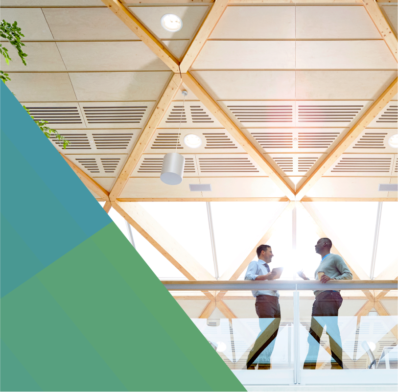 photo of two men talking on a walkway in their open office. The viewer is looking up at them in an angle that allows them to see the ceiling with its lights, wooden beams, and windows.